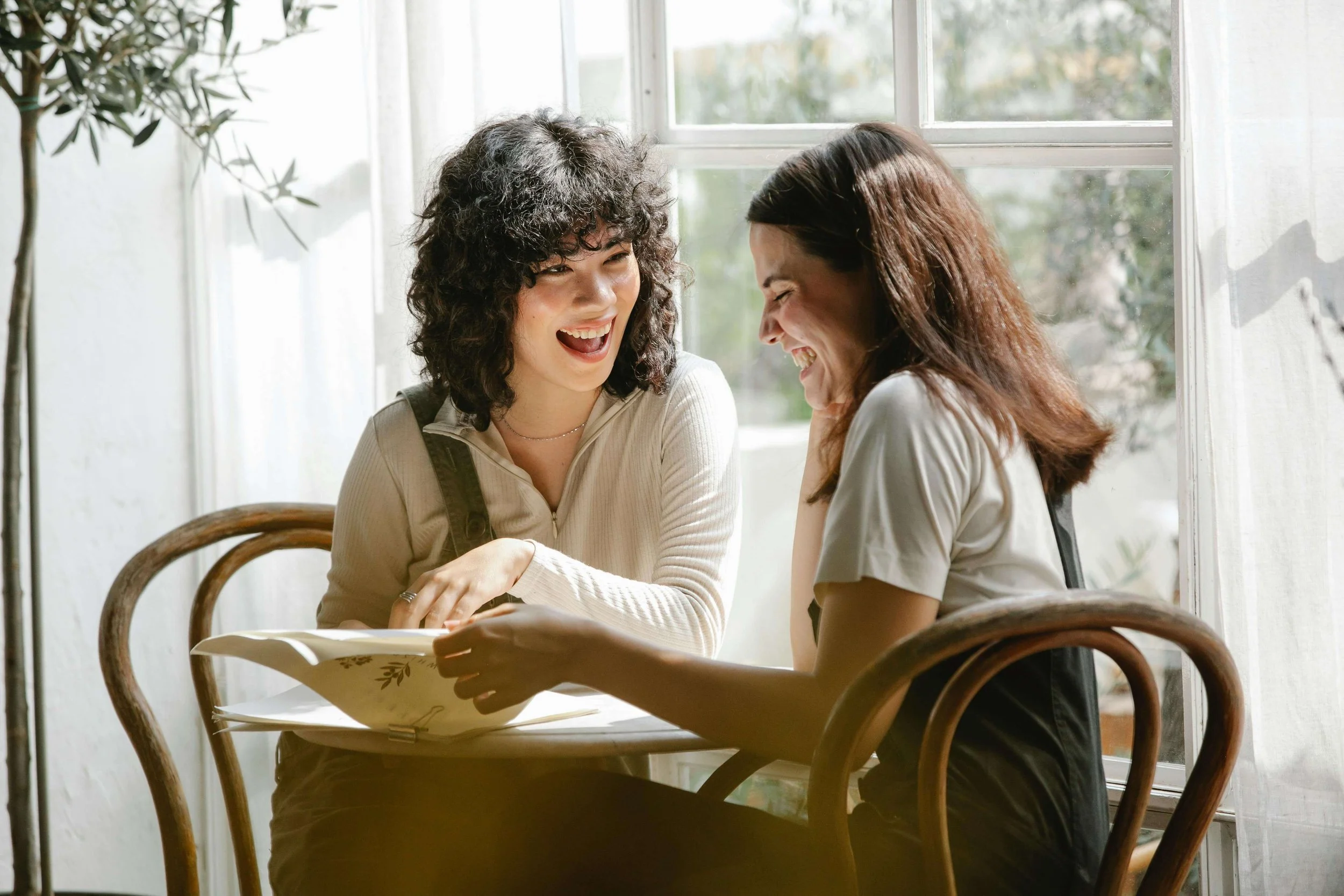 Two biracial friends smiling while sitting at a cafe table. Discover how ERP for relationship OCD in Boston, MA can help you navigate reassurance seeking & constant checking.