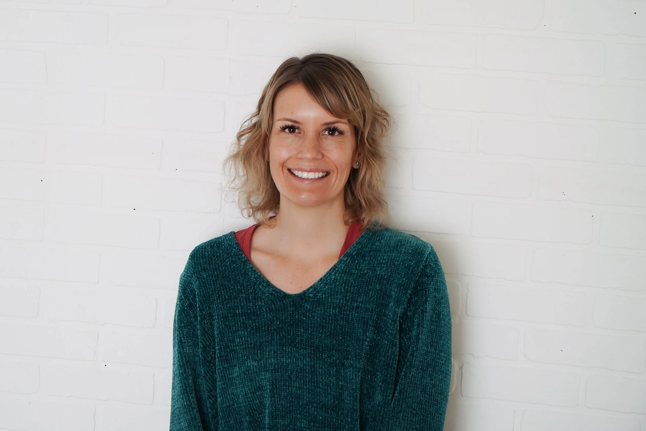 Jen Lescher smiling in front of a white brick wall. Jen offers OCD counseling in San Diego, CA for anyone struggling with obsessive thoughts, ROCD & more.