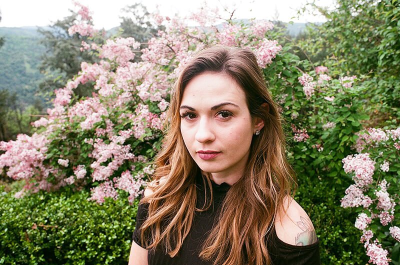 A woman softly smiling standing in front of small pretty pink flowers. Learn how an OCD therapist in Boston, MA can help you stop overthinking. Start your healing journey today.